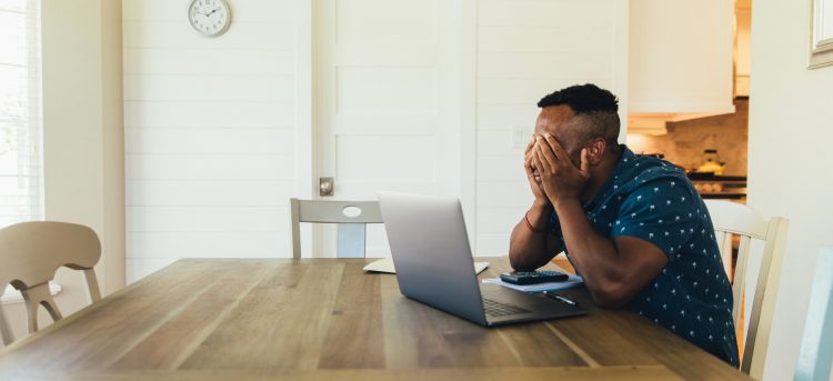 A 30 year old man works on his finances at his kitchen table. He is working on his bank account numbers and preparing to pay his taxes. Times are tough and making ends meet is difficult.